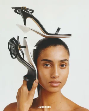Model Imaan Hammam poses with stacked black and white Jacquemus strappy kitten-heel sandals balancing on her head on a white backdrop.
