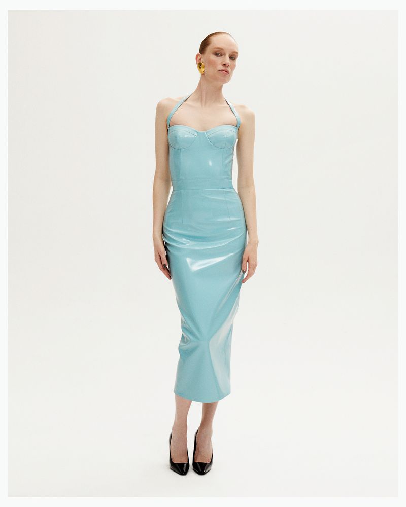 Elegant woman in a shiny aqua halter dress with black heels posing in a white photo studio with soft lighting and wearing yellow earrings.