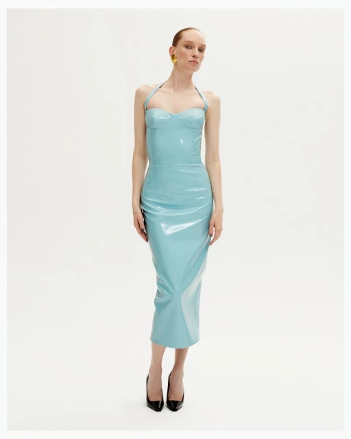 Elegant woman in a shiny aqua halter dress with black heels posing in a white photo studio with soft lighting and wearing yellow earrings.