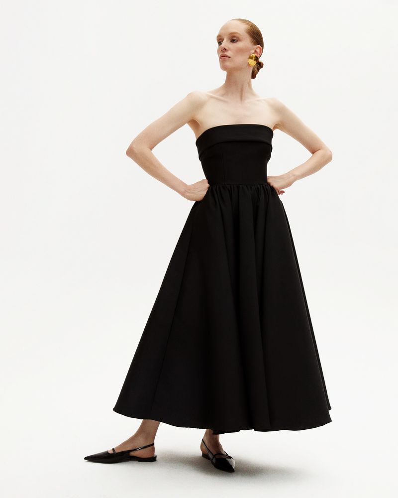 Elegant woman in black strapless dress and flats poses against a white backdrop in a fashion editorial shot.