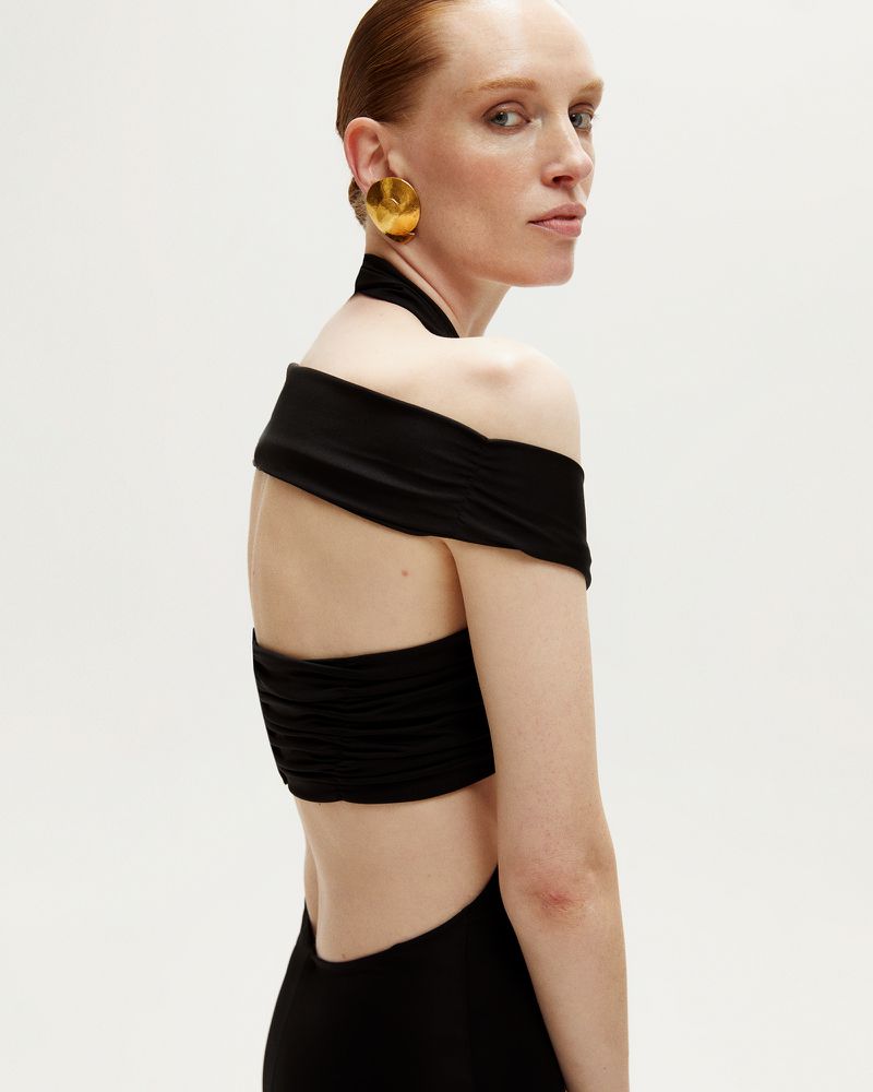 Adult female with red hair poses in off-shoulder black halter dress and gold earrings against a white backdrop in a studio shot.
