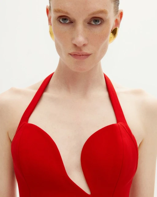 Close-up of a woman with a serious expression wearing a red halter top and gold earrings against a white background. Beauty shot.