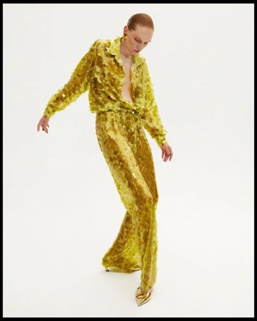 A slender adult woman in yellow sequined shirt and pantsuit strikes a fashion pose against a clean white backdrop, complemented by gold heels.
