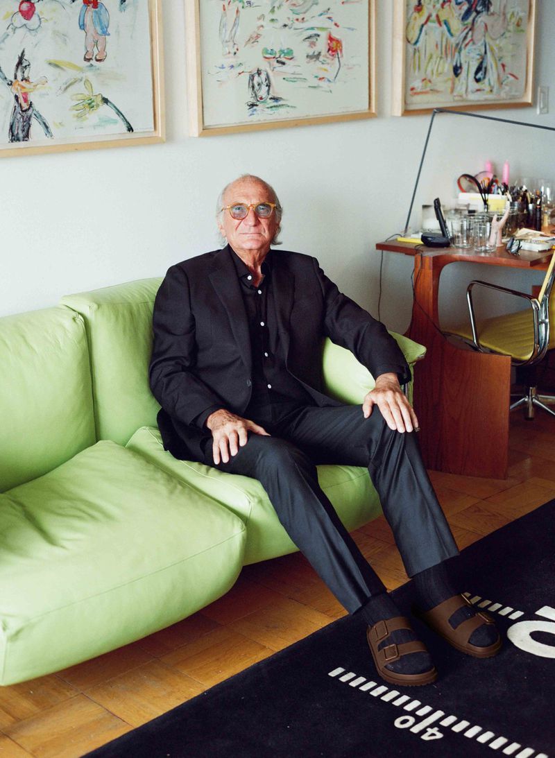 Elderly man in black suit sits on a green couch in a bright room with artwork and a desk. He wears sandals on a patterned rug.