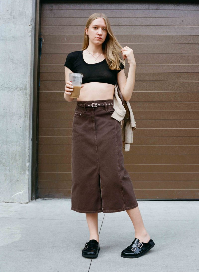 A young woman poses outdoors, wearing a black crop top, brown midi skirt, and black clogs, holding a drink against a brown metal door.
