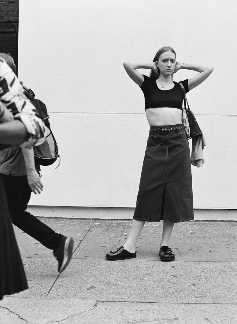 A young woman in a black crop top and denim skirt stands on a sidewalk next to a wall in a black and white urban setting.
