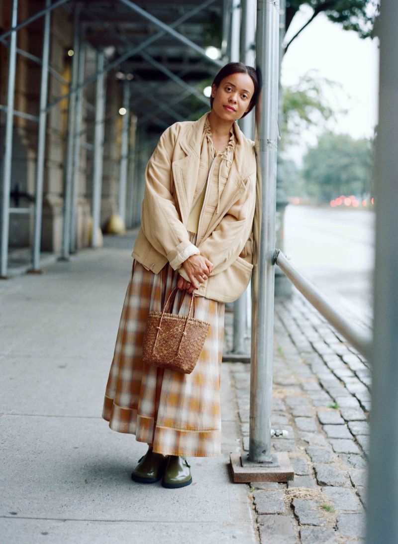Woman in plaid skirt and blazer stands outdoors, leaning on scaffolding with a woven bag, on a city street with trees and buildings.