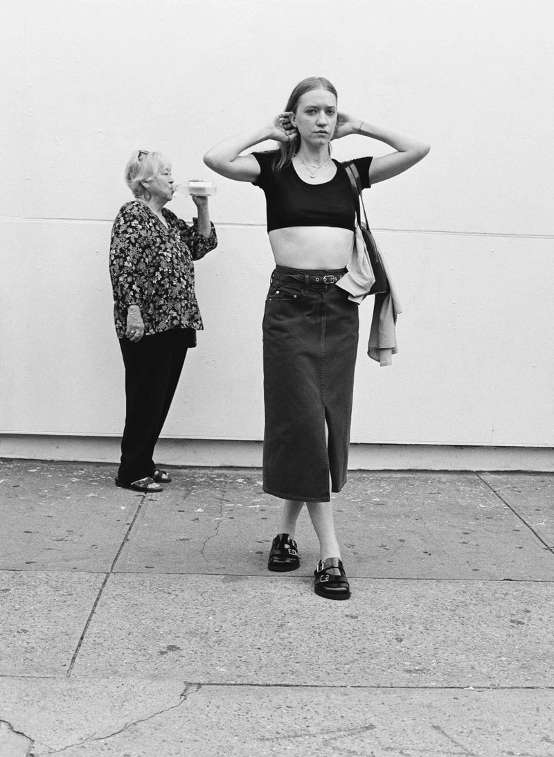 Black and white shot of a young woman in a black top and long denim skirt standing next to an elderly woman drinking from a cup.