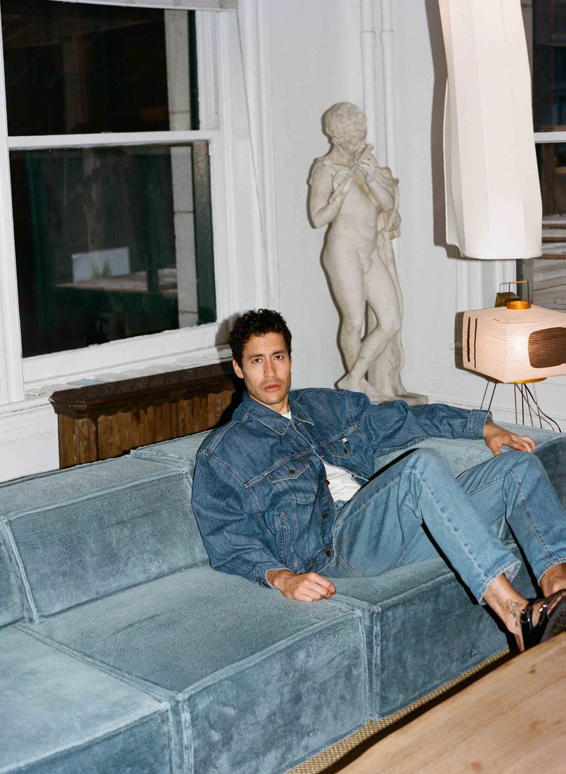 Adult male in denim jacket and jeans lounging on a blue couch indoors, with a statue and modern lamp in the background, medium shot.