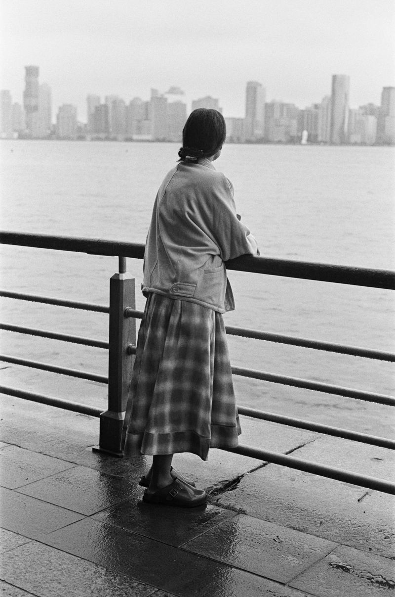 Monochrome shot of a woman in a plaid skirt gazing out at a distant city skyline from a pier, with a calm body of water in between.