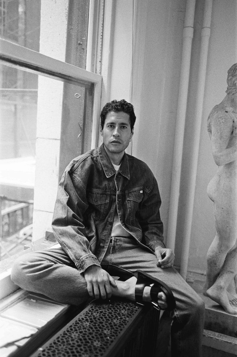 Monochrome portrait of a man sitting cross-legged on a windowsill with a denim jacket and jeans, statue in the background.