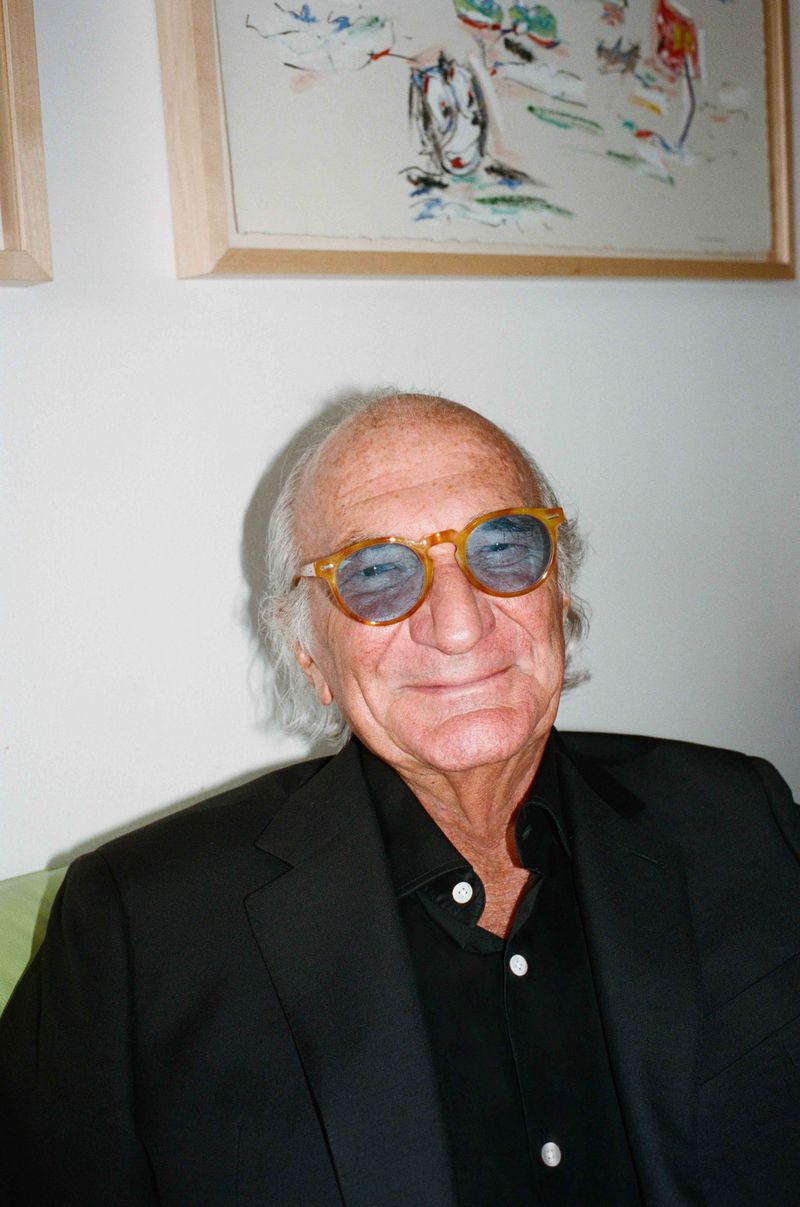 Portrait of an older man with white hair, wearing a black suit and yellow-tinted glasses, smiling indoors with art on the wall.