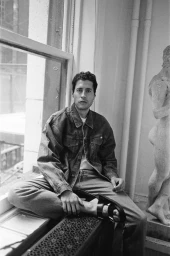 Black and white photo of a man wearing a denim jacket, jeans, and black sandals seated on a radiator by a window next to a statue.