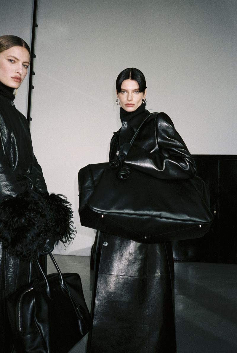 Two fashion models in black leather coats with a large black leather bag in a neutral indoor setting with neutral background.