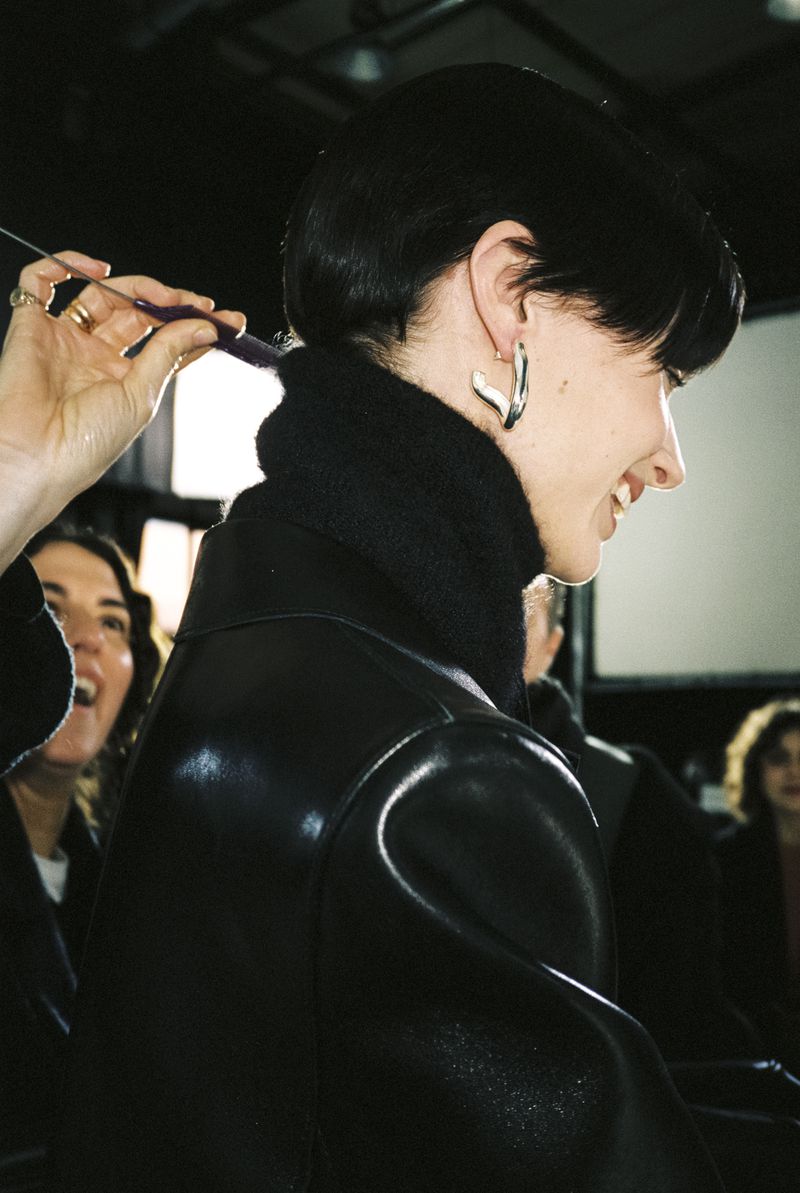 Model Mila van Eeten in profile wearing a black leather jacket and silver earring while having her hair styled backstage.