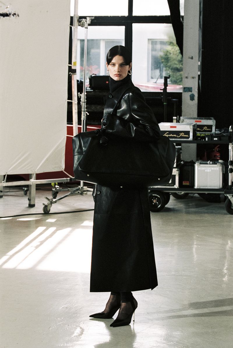 A woman in a black coat and heels poses with a large bag in a studio setting with lighting equipment and a white backdrop.