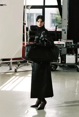 A woman in a black coat and heels poses with a large bag in a studio setting with lighting equipment and a white backdrop.