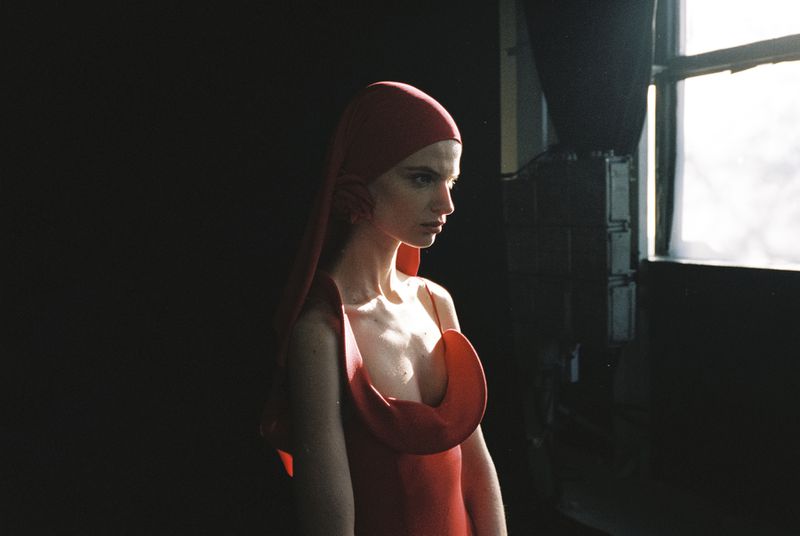 Woman in red headscarf and dress posing in dark room with window. Side view fashion shot with natural light, dramatic shadow.