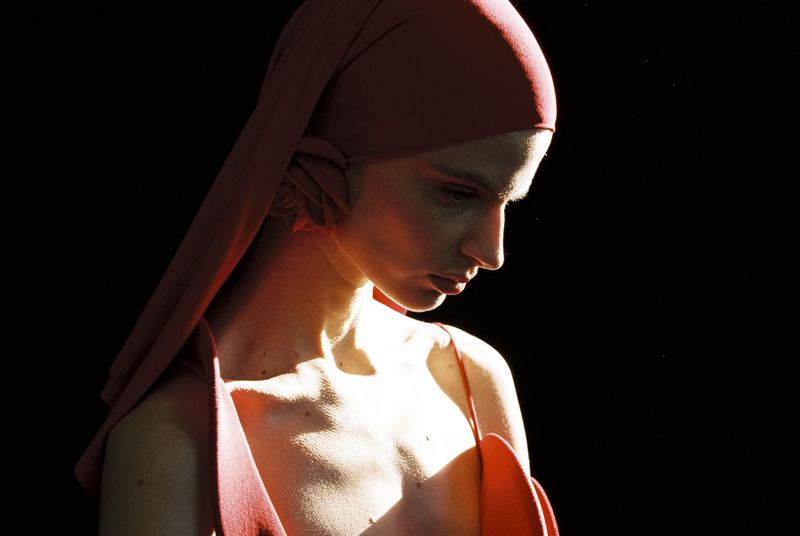 Portrait of a pale-skinned woman with a red headscarf looking down, bathed in light against a dark background, fashion beauty shot.