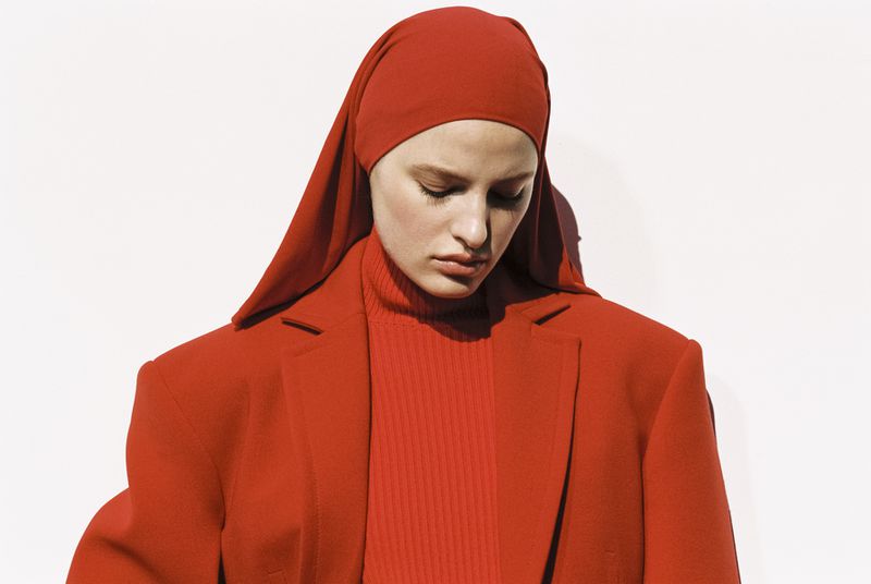 Adult female fashion model in red headscarf, turtleneck sweater, and blazer poses against a white backdrop in studio lighting.