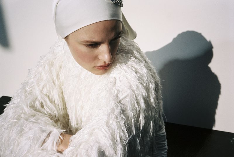 Woman with pensive expression in white textured coat and cap against a white background, capturing a minimalist fashion moment.