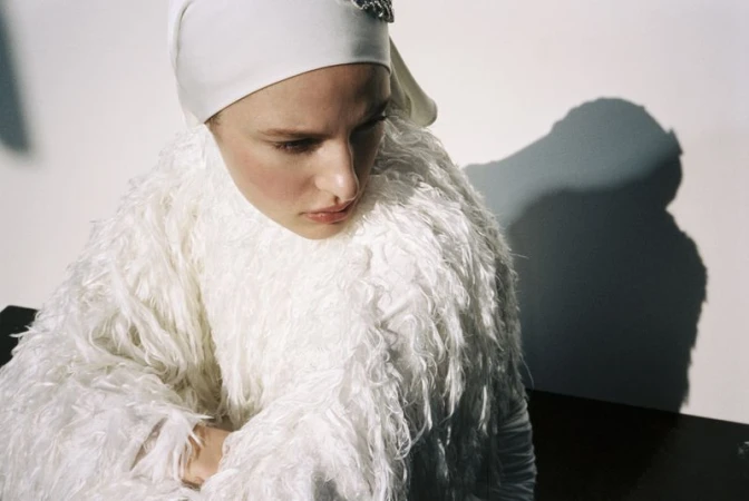 Woman with pensive expression in white textured coat and cap against a white background, capturing a minimalist fashion moment.