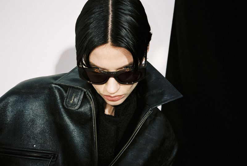 Close-up of a dark-haired fashion model with sunglasses, wearing a black leather jacket, set against a white and black background.