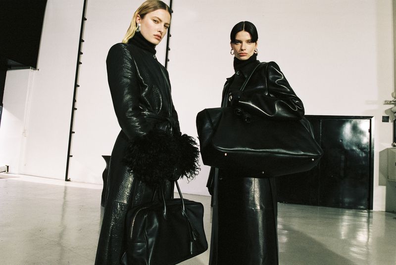 Two adult women in black leather coats pose with handbags in a photo studio with white walls and diffused lighting, fashion editorial.
