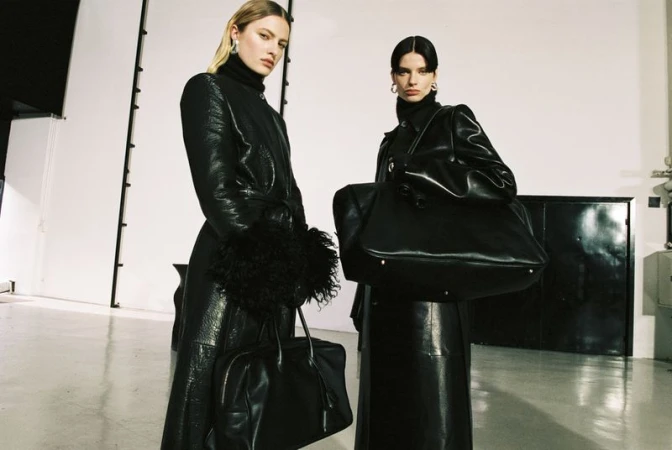 Two adult women in black leather coats pose with handbags in a photo studio with white walls and diffused lighting, fashion editorial.