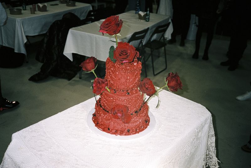 Two-tiered red cake decorated with red roses, set on a white tablecloth in an indoor event space with tables and chairs.
