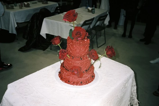 Two-tiered red cake decorated with red roses, set on a white tablecloth in an indoor event space with tables and chairs.