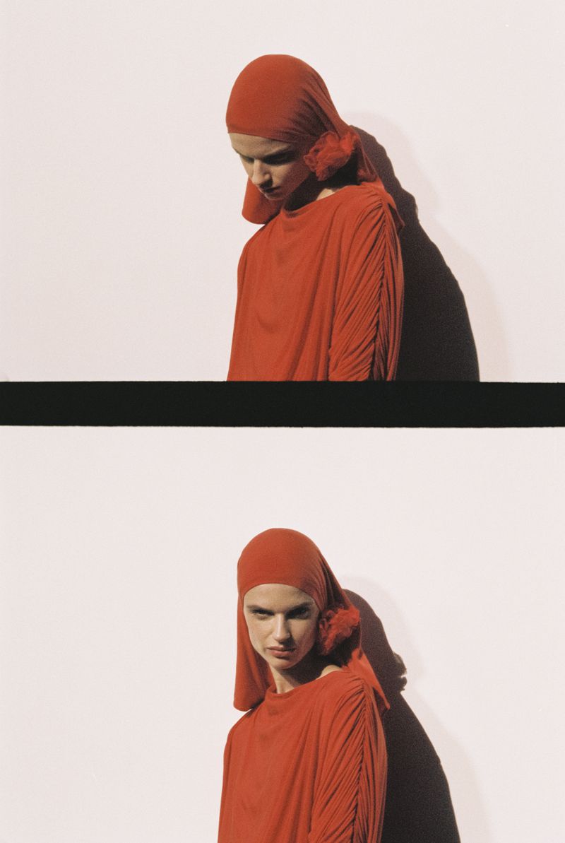 A woman wears a red hooded dress in a studio shot; shadows add depth to the composition against a white backdrop.