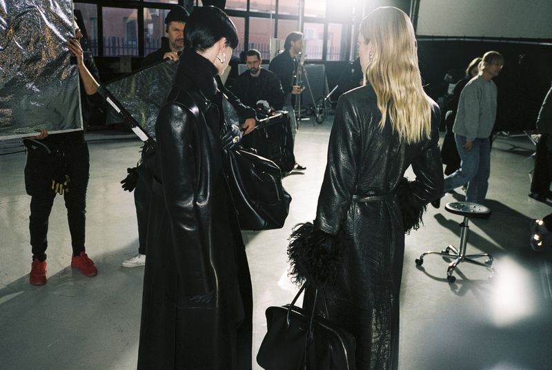 Two fashion models in leather coats on a photo studio set with crew in background, illuminated by studio lighting and a silver reflector.