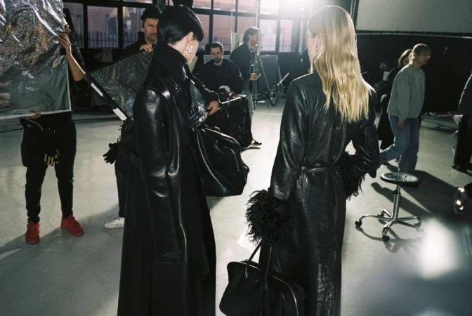 Two fashion models in leather coats on a photo studio set with crew in background, illuminated by studio lighting and a silver reflector.