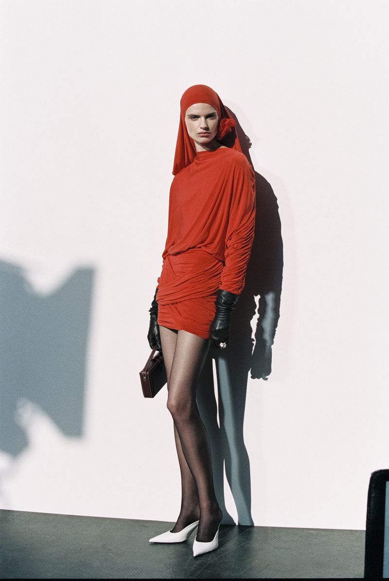 Fashion shot of a female model in a red dress and headscarf with black gloves and tights, holding a purse against a neutral background.
