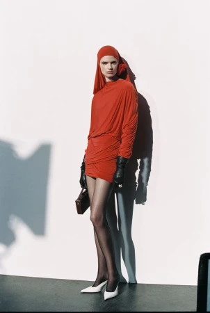 Fashion shot of a female model in a red dress and headscarf with black gloves and tights, holding a purse against a neutral background.