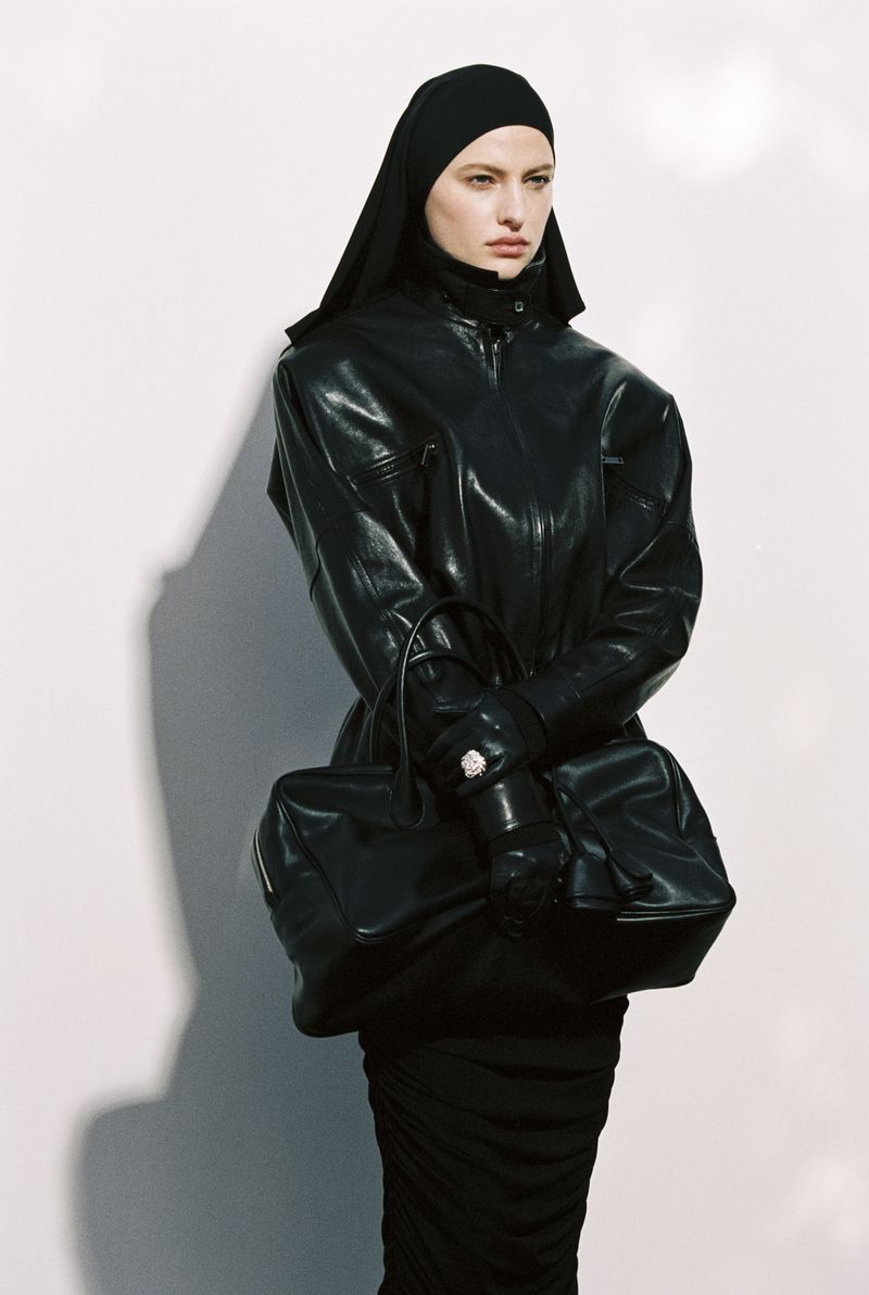 Woman in black leather jacket, head covering, and fitted dress, carrying a structured handbag against a neutral wall.