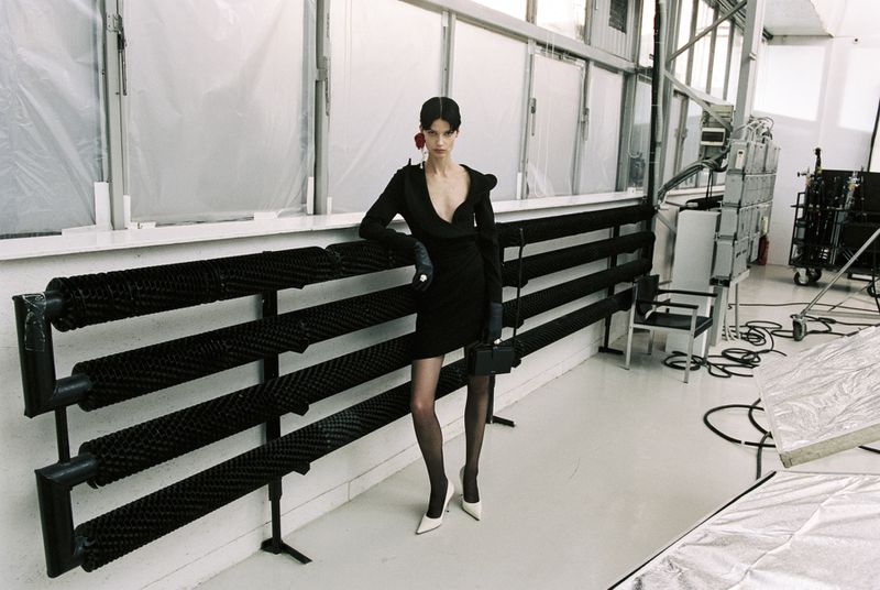 A model in a black dress poses in a studio with white floors, black radiator, and diffused natural light showcasing fashion elegance.