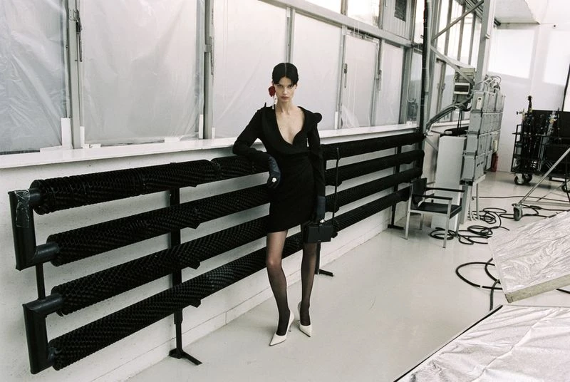 A model in a black dress poses in a studio with white floors, black radiator, and diffused natural light showcasing fashion elegance.