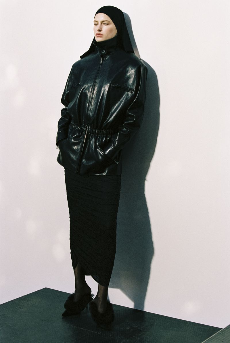 Woman in a black head covering and leather jacket poses against a white wall. She wears a fitted midi skirt and slipper-like shoes.