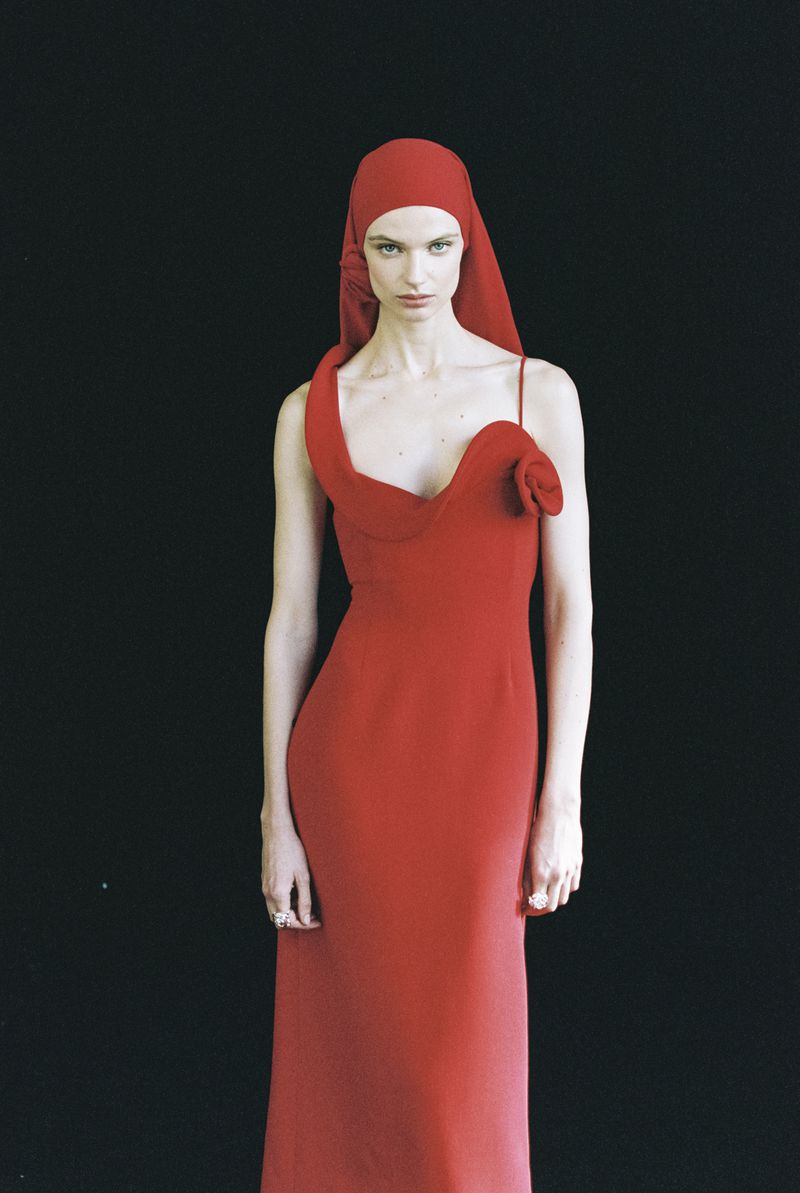 A slender woman in a red gown and matching headscarf poses against a black backdrop, showcasing a refined and elegant fashion aesthetic.