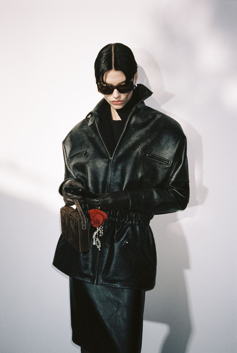 Woman in leather jacket, gloves, and skirt holding a purse with a red rose against a white backdrop in a studio setting. Fashion shot.