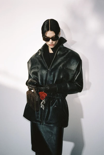 Woman in leather jacket, gloves, and skirt holding a purse with a red rose against a white backdrop in a studio setting. Fashion shot.