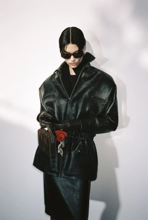 Woman in leather jacket, gloves, and skirt holding a purse with a red rose against a white backdrop in a studio setting. Fashion shot.