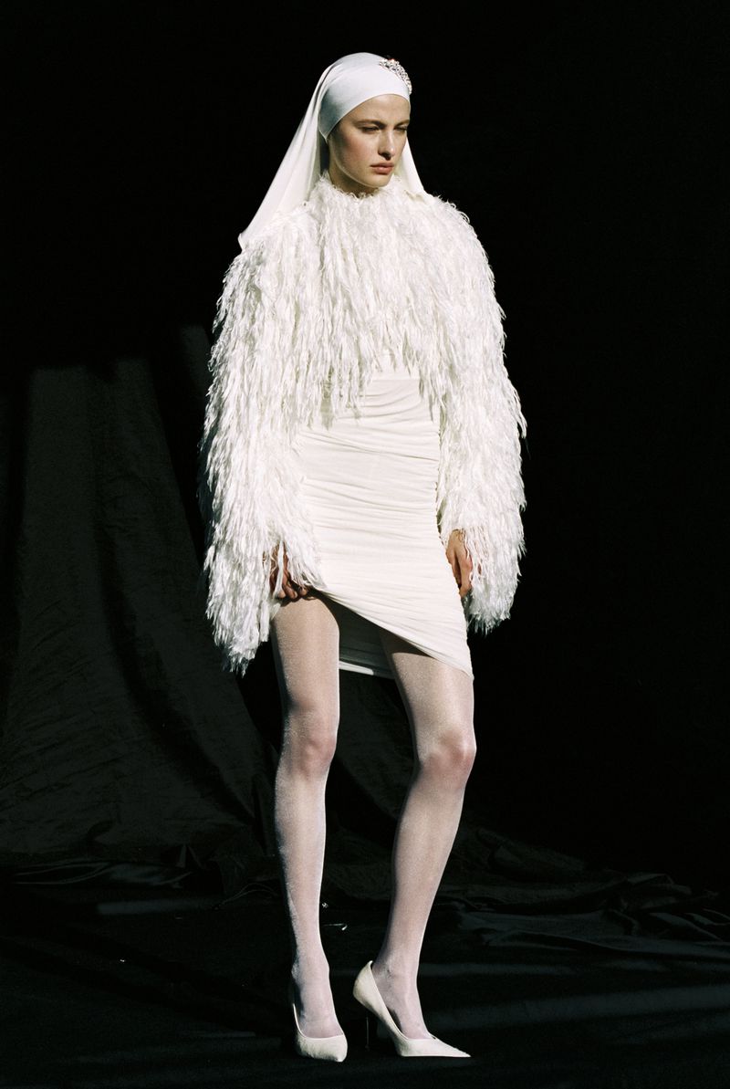 A fashion model in an all-white outfit with a fringed jacket and head covering poses in a dimly lit studio.