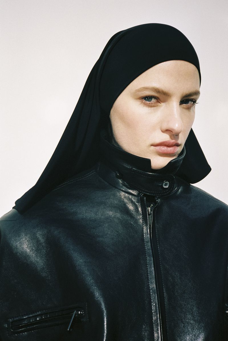 Model wearing a black leather moto jacket with a high collar and a matching black headscarf against a plain white studio backdrop.