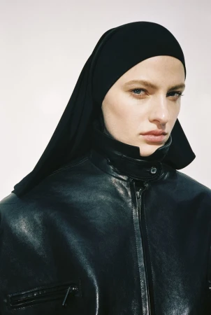 Model wearing a black leather moto jacket with a high collar and a matching black headscarf against a plain white studio backdrop.