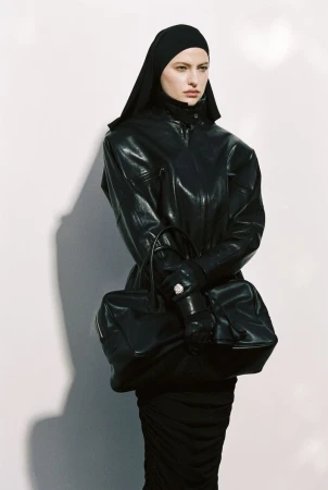 Model wearing dark leather jacket and head covering holding black handbag against white seamless background
