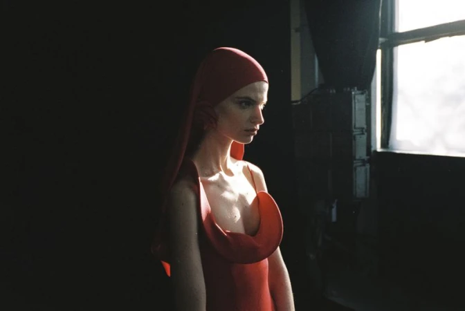 Model wearing a red hooded dress with a sculptural neckline in a moody studio setting by Vitali Gelwich.