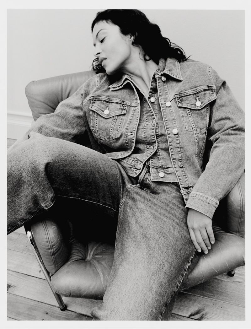 Model seated in an armchair wearing a denim jacket and jeans against a plain background